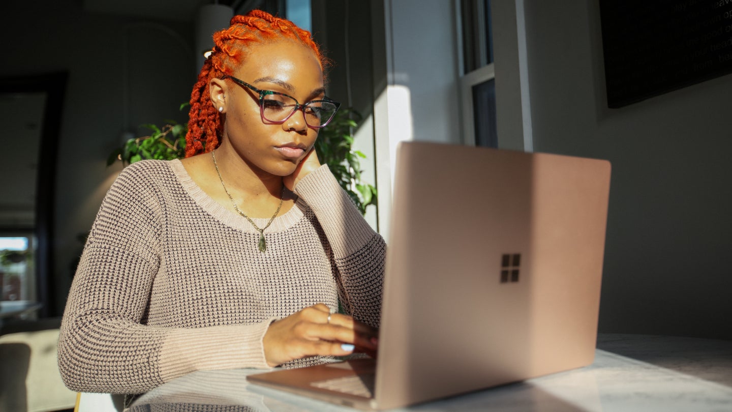 A Black woman with orange, braided hair sitting in front of a silver Surface laptop, possibly using Microsoft PowerToys.
