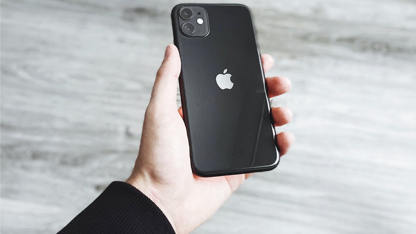 A person holding a black iPhone 11 over a gray wood floor.