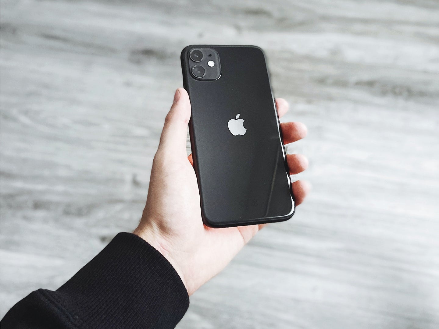 A person holding a black iPhone 11 over a gray wood floor.