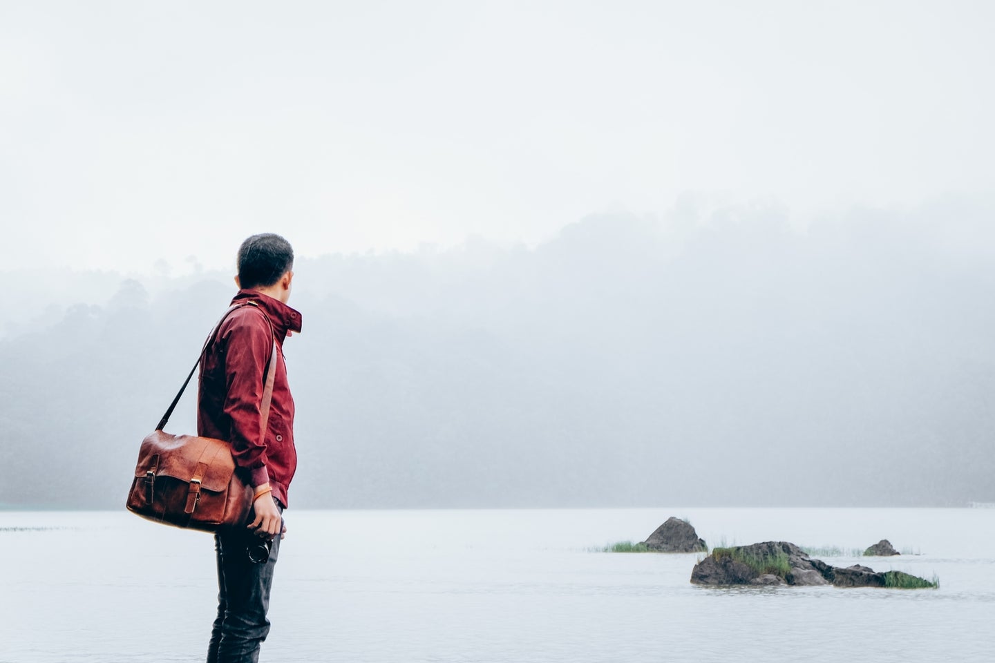 a man in a red jacket and jeans carrying a brown leather messenger bag and staring out at gray water and some rocks covered in fog on a cloudy day