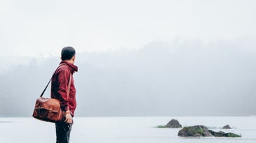 a man in a red jacket and jeans carrying a brown leather messenger bag and staring out at gray water and some rocks covered in fog on a cloudy day
