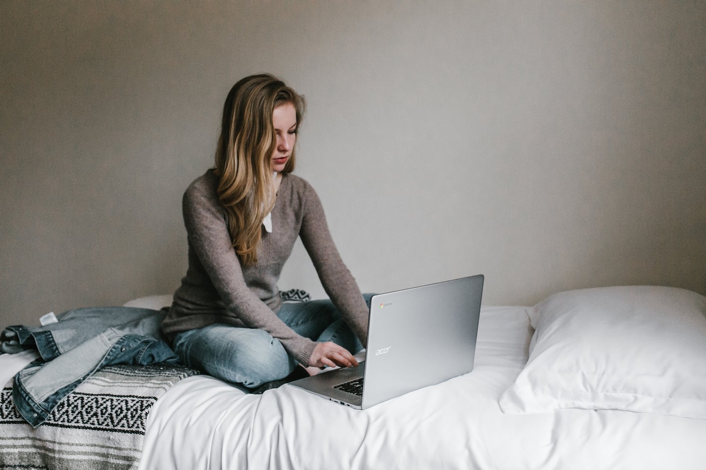 a woman sitting on a bed with white blankets, using a Chromebook