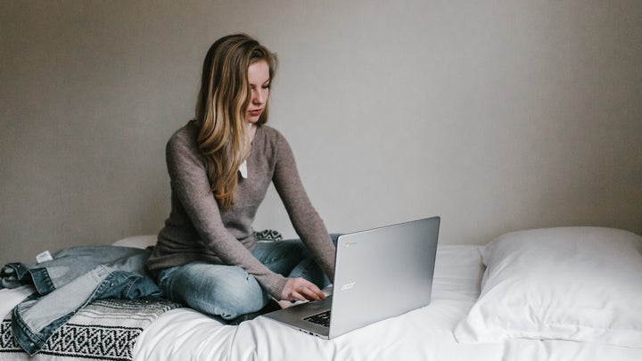 a woman sitting on a bed with white blankets, using a Chromebook