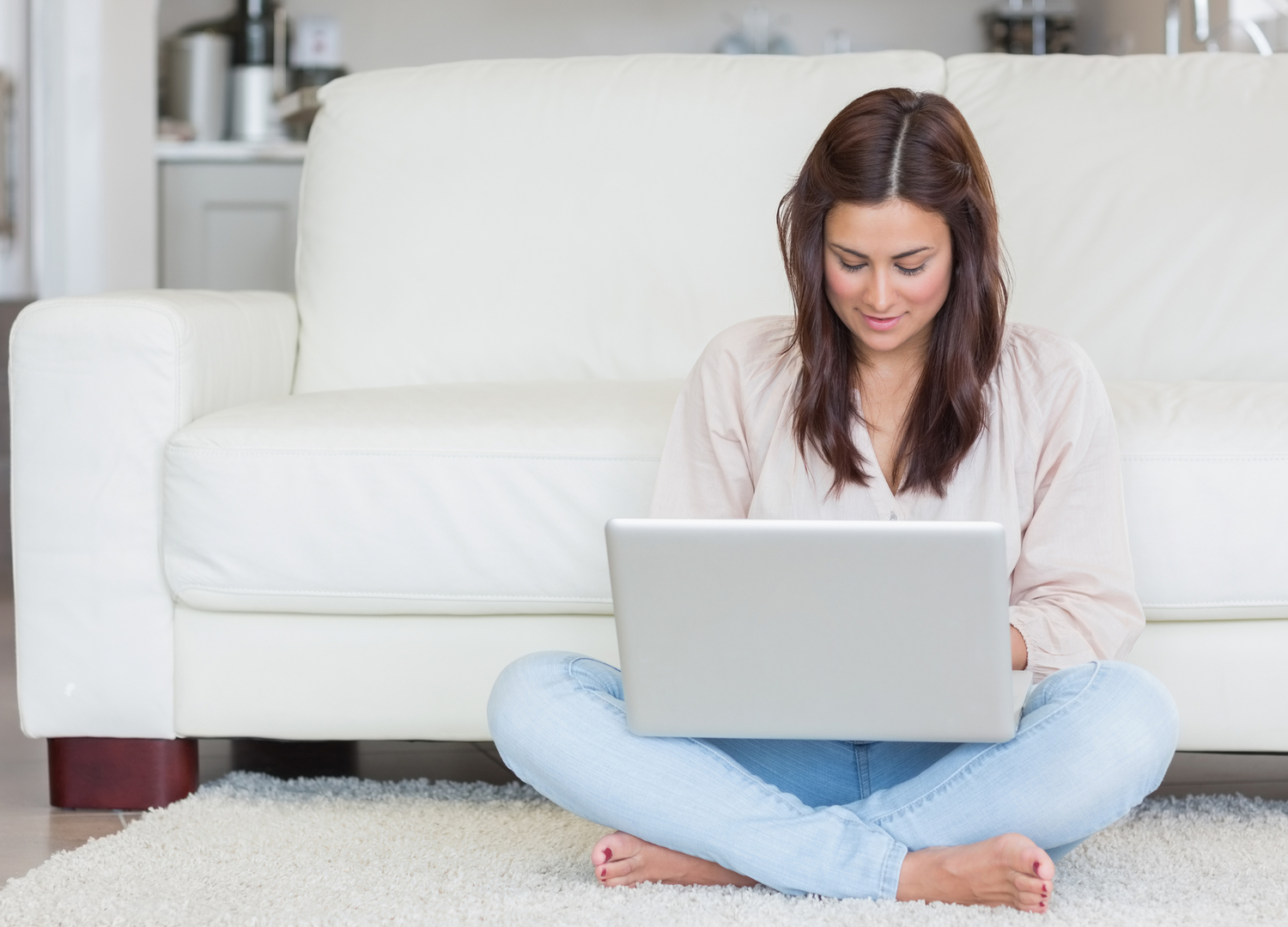A brunette woman wearing a light pink shirt and light blue jeans sitting on a white carpet in front of a white couch and typing on her new silver laptop.