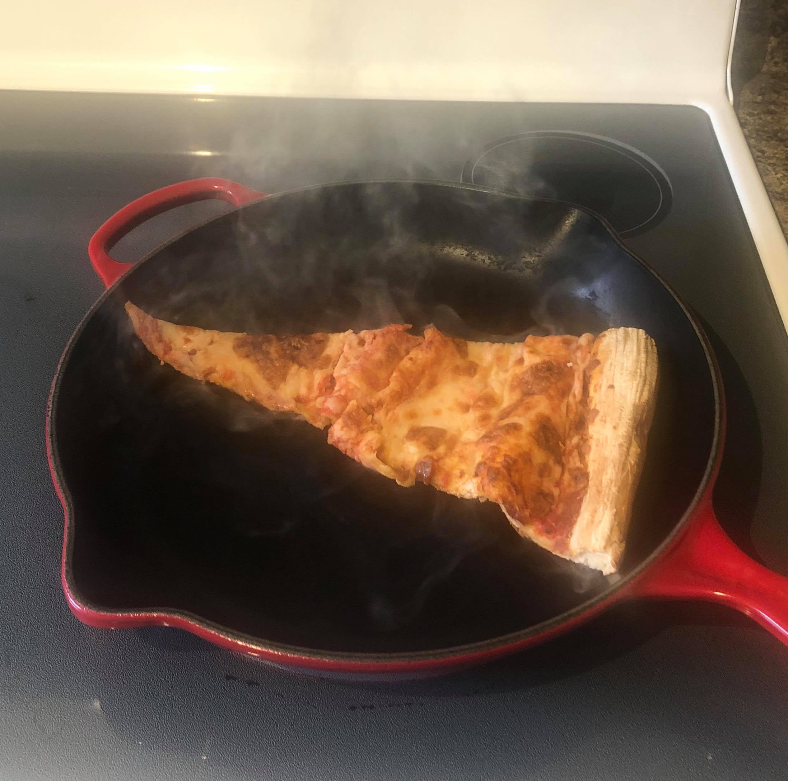 A slice of leftover cheese pizza burning on a cast-iron pan on an electric stove.
