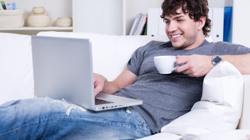 a man on his white couch with a cup of tea or coffee, smiling while using his laptop