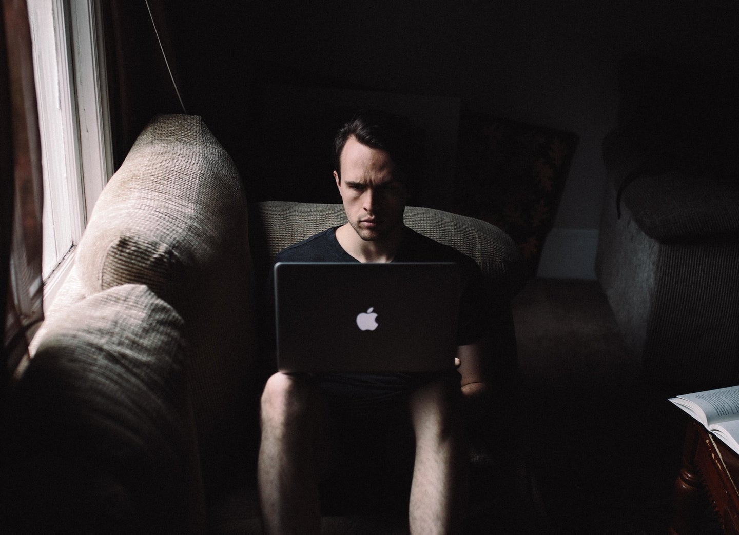 A man sitting solemnly on a couch in the dark with his Apple Mac laptop