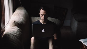 A man sitting solemnly on a couch in the dark with his Apple Mac laptop