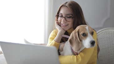 A woman smiling at her computer and holding a dog