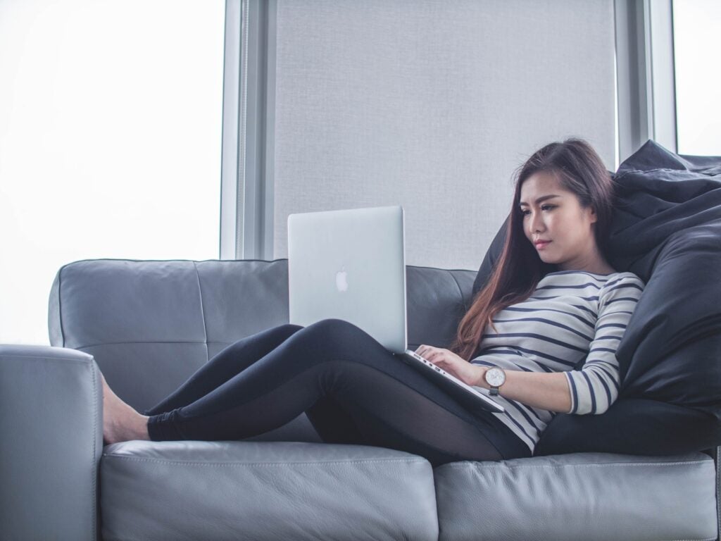 a woman sitting on a couch with a lot of pillows while using an Apple Macbook laptop