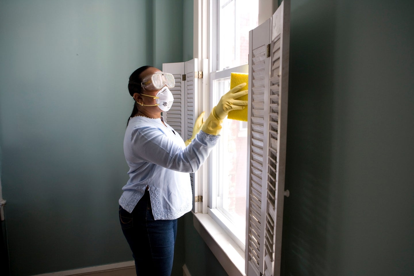 a woman wearing a face mask, goggles, and gloves while cleaning a window with a sponge