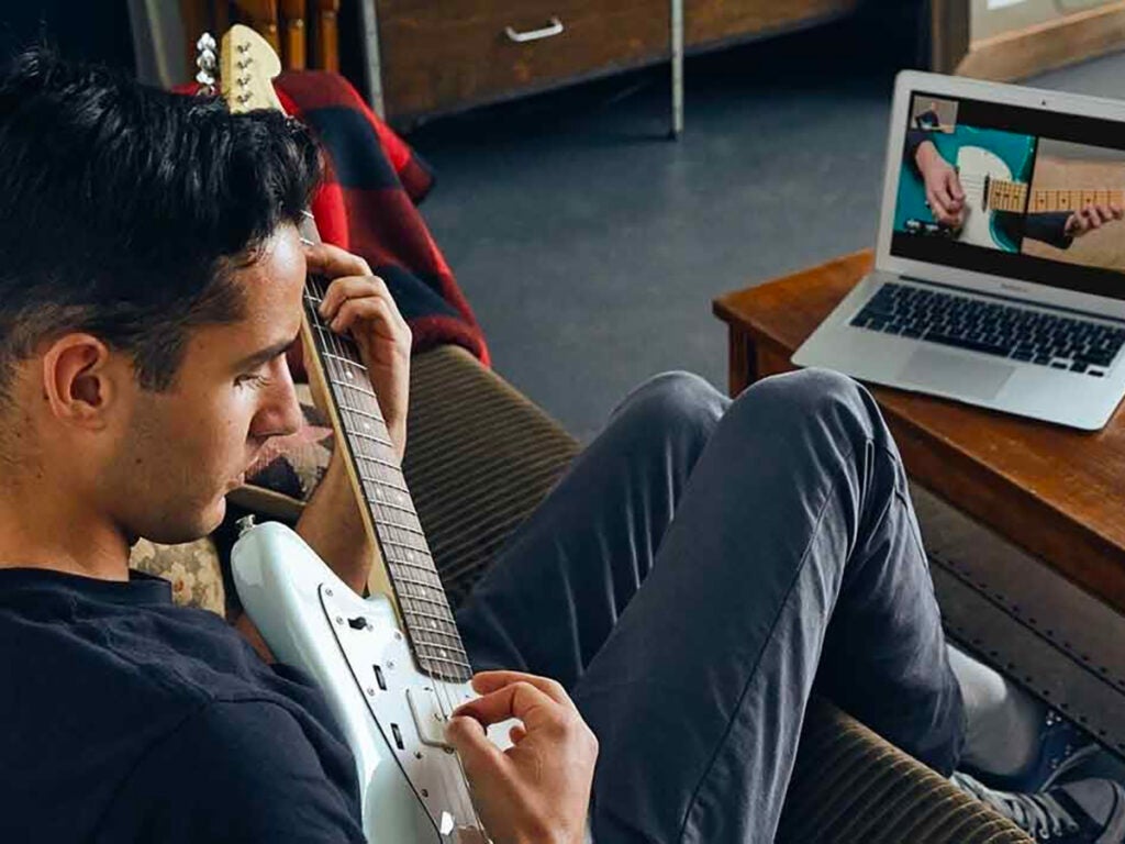a man sitting on a couch learning guitar from a tutorial on his laptop computer