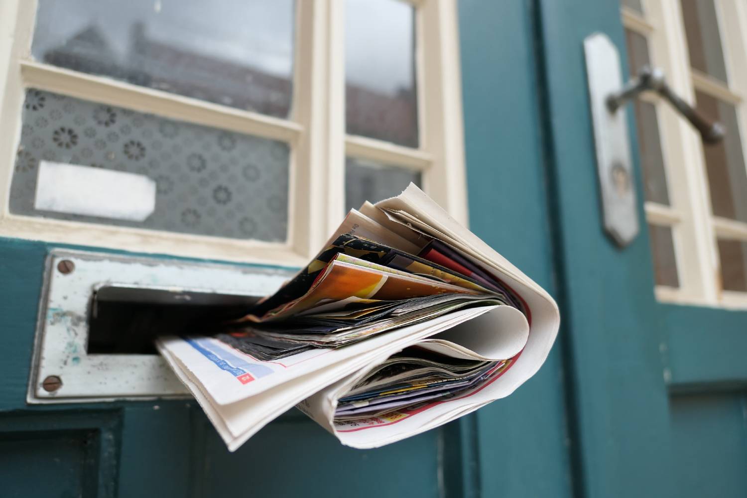 A magazine sticking out of a mail slot in a green door with glass window panes.