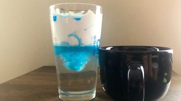 a photo of a person pouring colored water into a glass of shaving cream and water to demonstrate rainfall with a science experiment project creating a storm in a glass