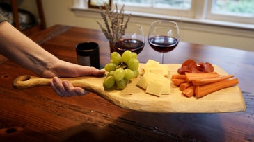 a person holding a handmade DIY cutting board with cheese, grapes, carrots, and strawberries on it