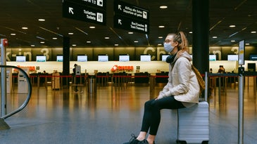 woman wearing mask sitting on suitcase in airport