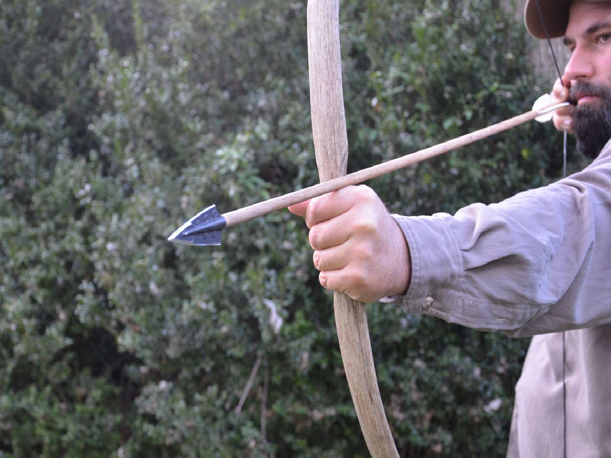 A man with a beard draws back on a hand-made bow and arrow set.
