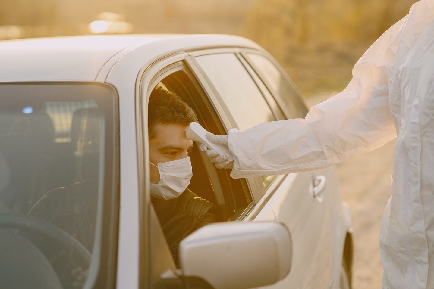a man in a car wearing a mask is having his temperature taken by someone wearing protective gloves