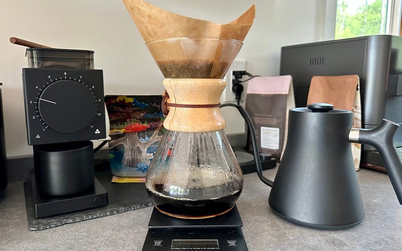 Chemex pour over coffee maker on a counter with coffee, grinder, and kettle