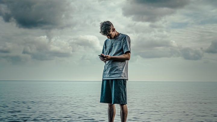 A person standing on a rock by the water under cloudy skies while looking at his phone—hopefully he has a good weather app.