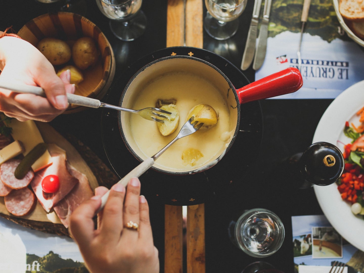 two people doing fondue with a bunch of food and using forks to dip potatoes in a pot of melted cheese