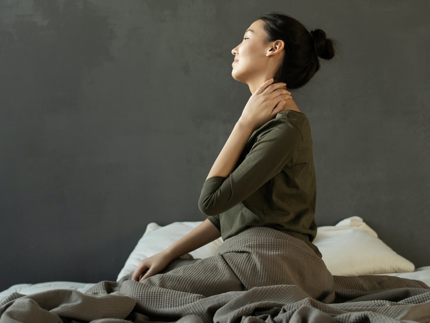 A black-haired woman with her hair in a bun, sitting in bed with neck pain.