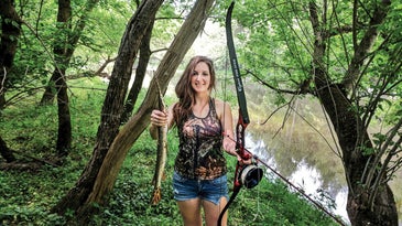 Woman holds a recurve bowfishing bow nocked with an arrow in one hand and a gar in the other hand while standing on a tree-lined creek bank.