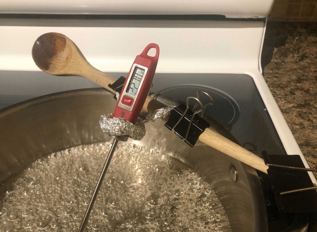 a digital thermometer measuring the temperature of melted sugar while attached to a wooden spoon with aluminum foil and binder clips