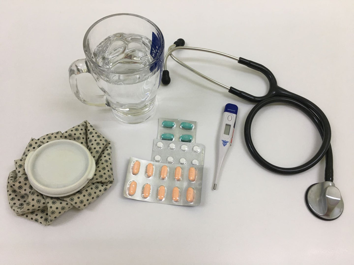 A collection of items used to ease flu symptoms: a glass of water, hot water bottle, allergy pills, a thermometer and a stethoscope