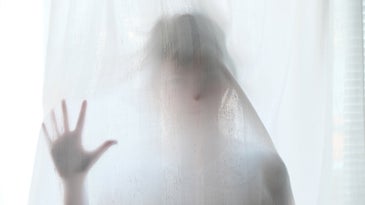 A person stands behind a white gauzy curtain in a spooky fashion