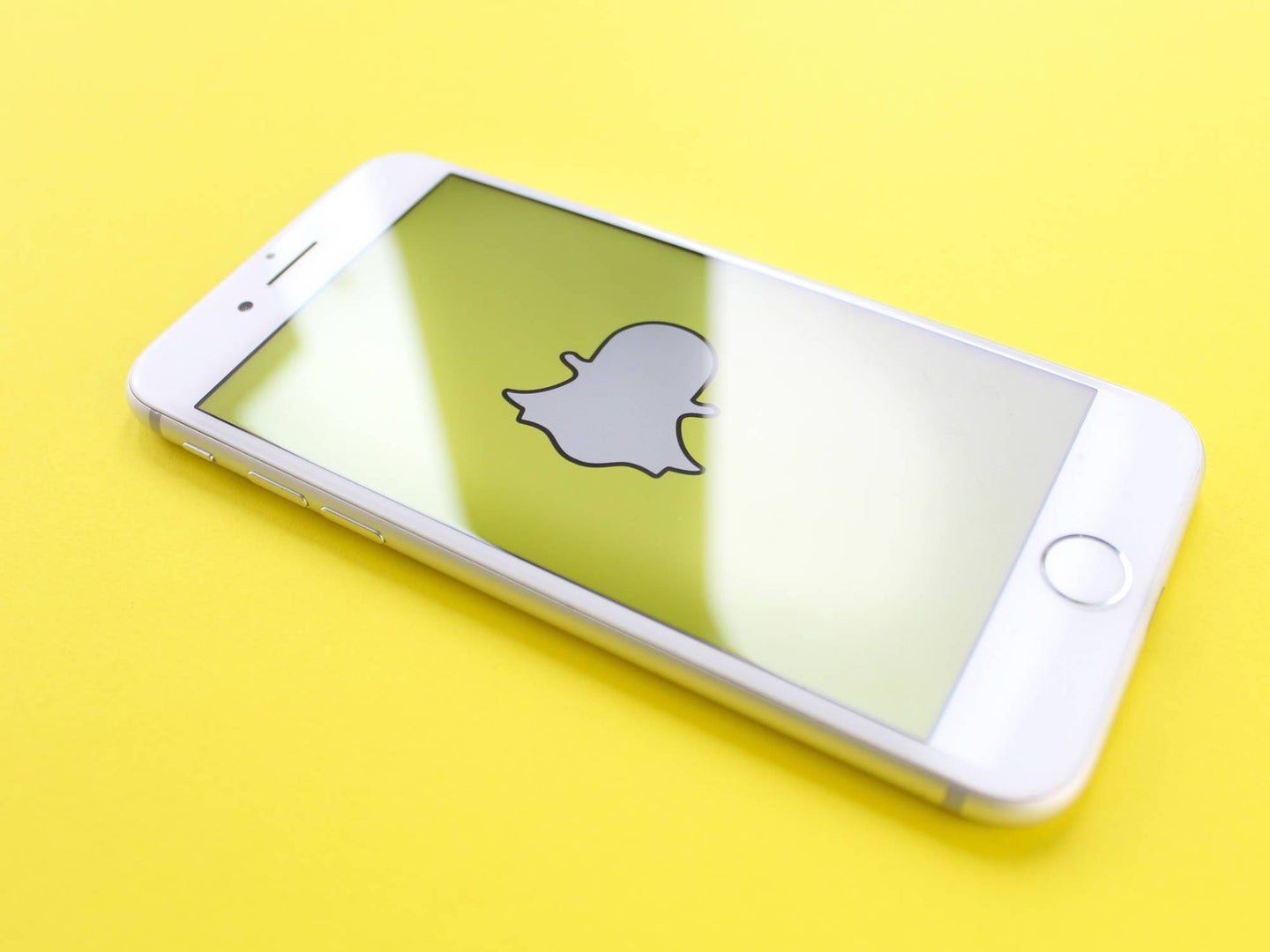 A white iPhone on a yellow background with the Snapchat ghost icon on the screen in landscape mode.