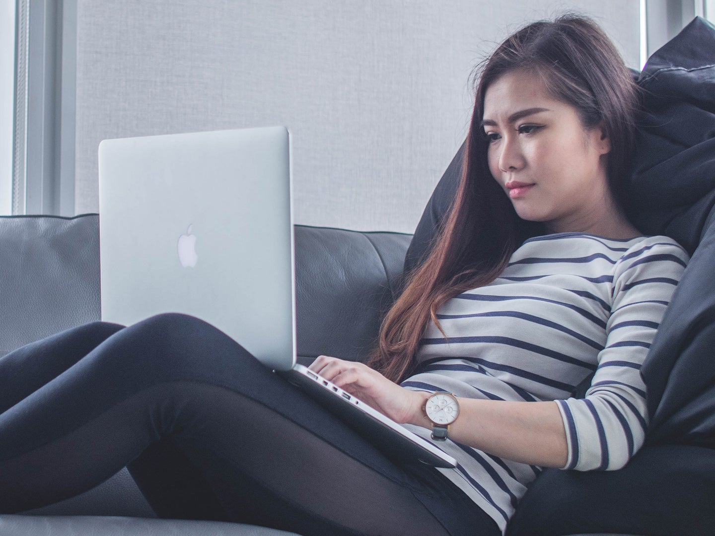 A woman lounging on a couch with a silver Macbook on her lap, possibly considering how she can create shortcuts for macOS.