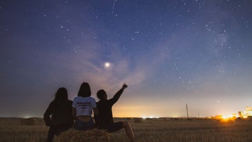 Three people watching the night sky while sitting on a hay bale in a field