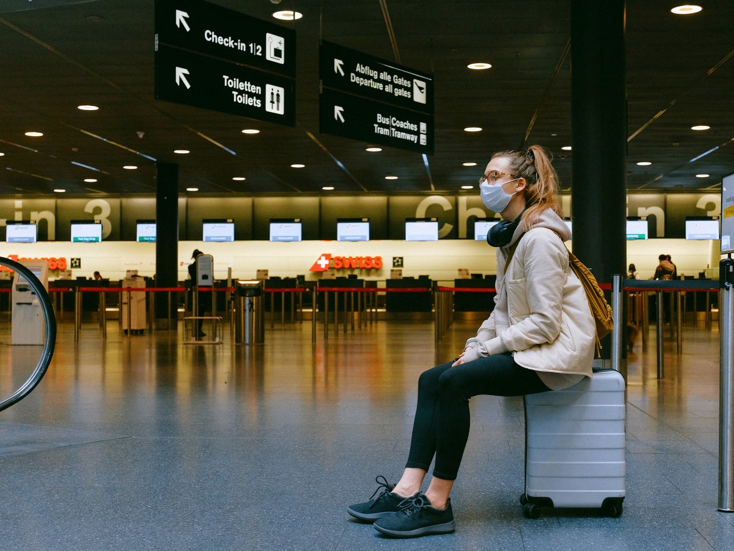 woman sitting on a suitcase in an airport wearing a face mask