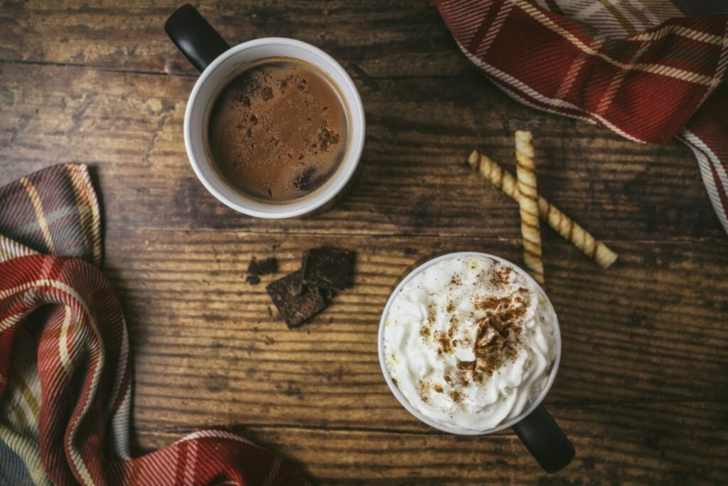 two mugs of hot cocoa, one with whipped cream and cinnamon on top, on a rustic looking wood table