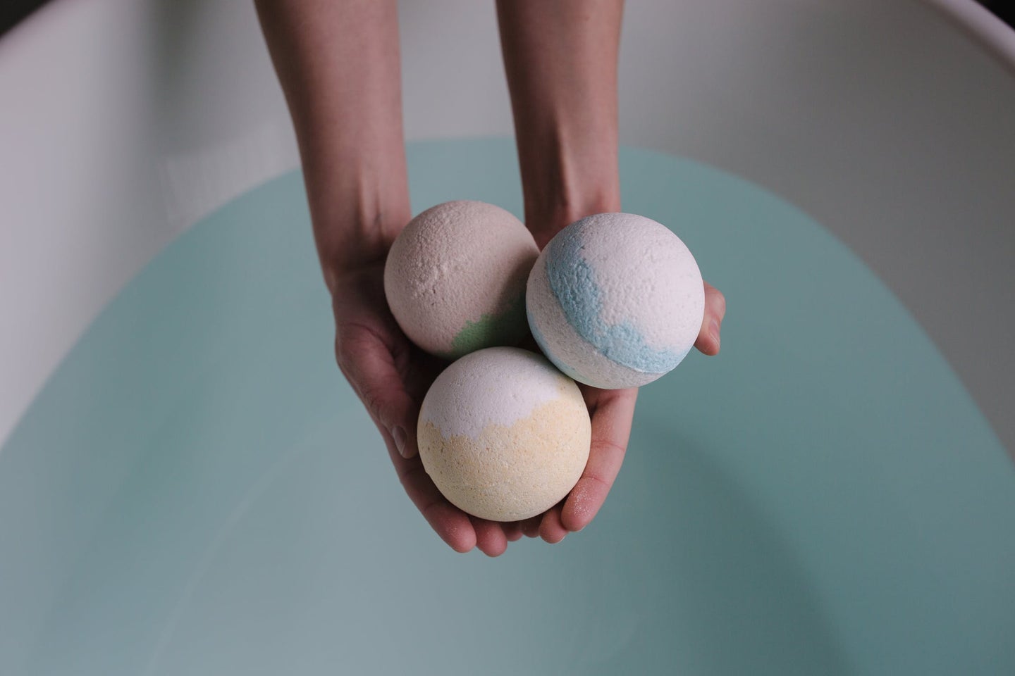 A pair of hands holds three homemade bath bombs over a bath tub full of water.
