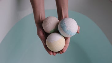 A woman holds three homemade bath bombs in her hand over a bath tub full of water.