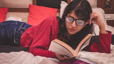 A person wearing glasses and laying on a bed while reading a book.