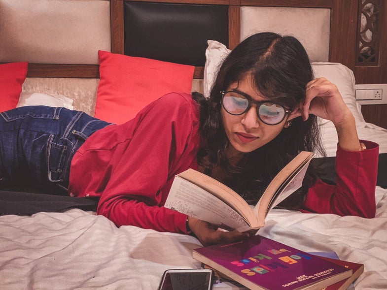 A person wearing glasses and laying on a bed while reading a book.