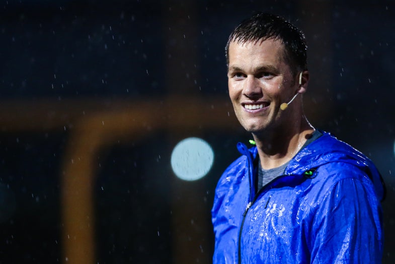NFL quarterback Tom Brady with an ear piece and in a blue rain jacket on a football field