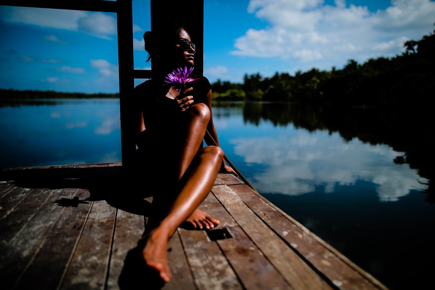 A person with dark brown skin tanning on a dock on a lake