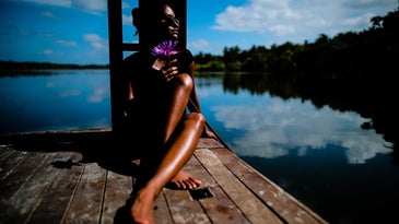 A person with dark brown skin tanning on a dock on a lake