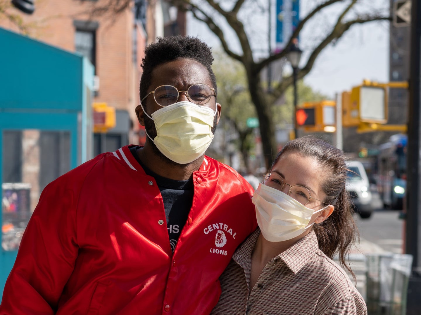 Two people wearing cloth face masks with their arms around each other on a New York City sidewalk on a sunny day.