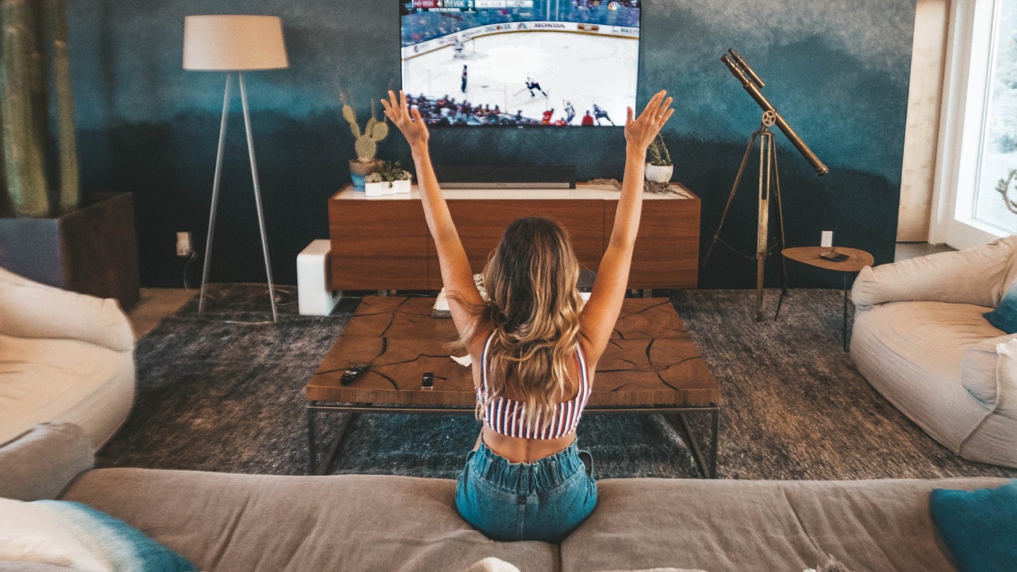 A woman cheering while sitting on a brown couch and watching a hockey game on a large flat-screen TV
