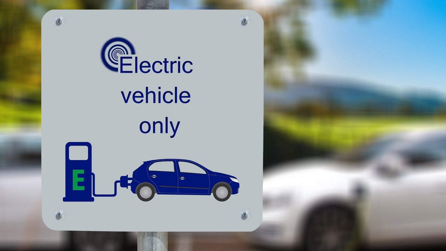 A sign saying the area is for electric vehicles only, with an image of an EV plugged into a charging station on it. Two white EVs are parked behind the sign.