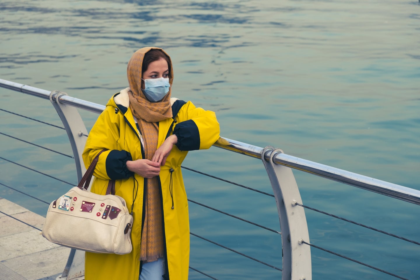A woman in mask, head scarf and yellow rain jacket stands along the water
