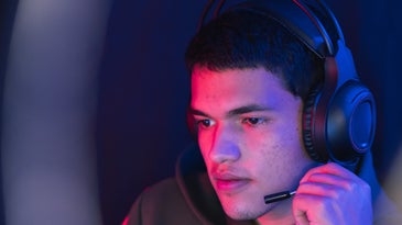 A young man under rd and purple light, wearing a computer headset and touching his microphone while looking at a screen.