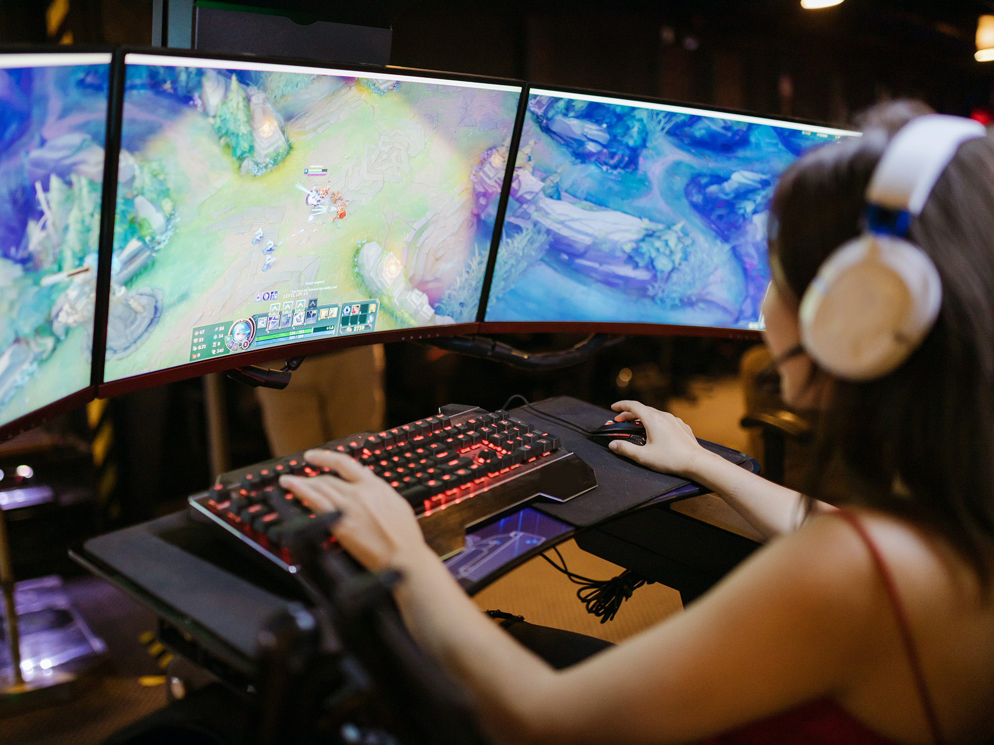 A woman wearing white headphones playing League of Legends on a triple-monitor setup.