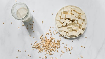 a bowl of tofu and a glass of soy milk on a white marble counter, with dried soybeans scattered around the surface