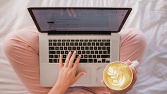 woman in pink pants sitting on bed with a computer and latte